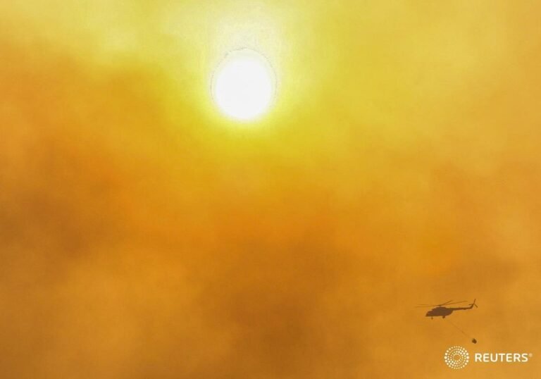 A firefighting helicopter flies through smoke from wildfires during sunset near